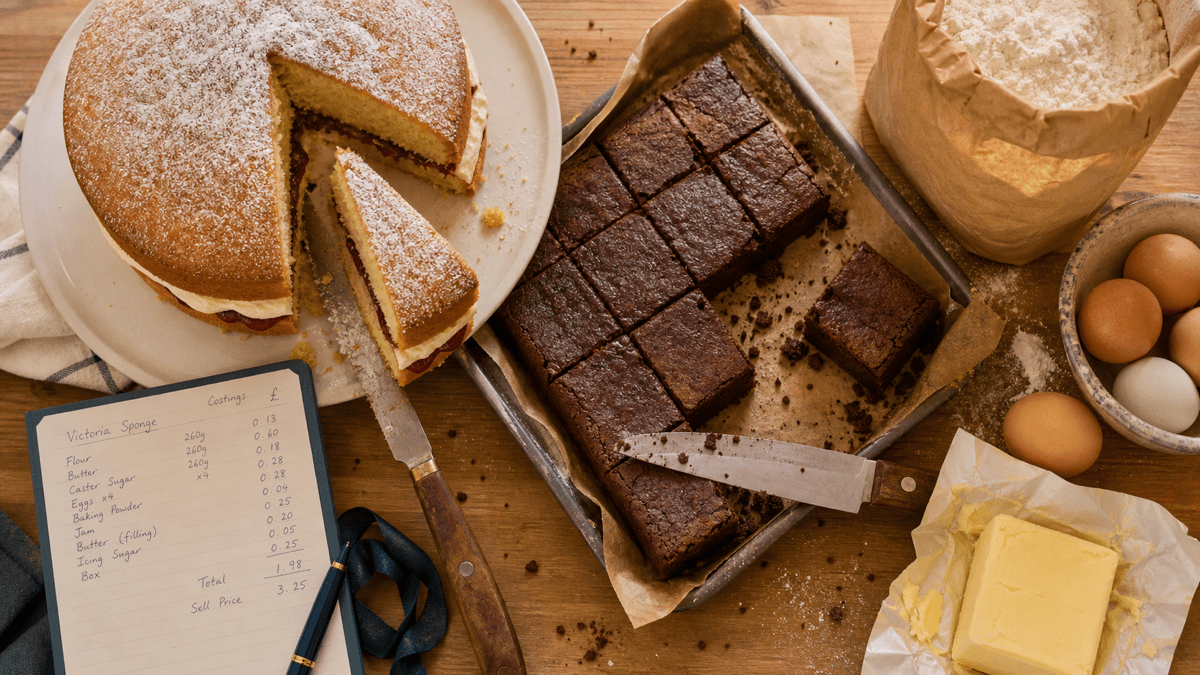A whole Victoria sponge and a tray of brownies being sliced on a bakery counter, surrounded by flour, butter, eggs and a recipe card with handwritten costings, warm golden light